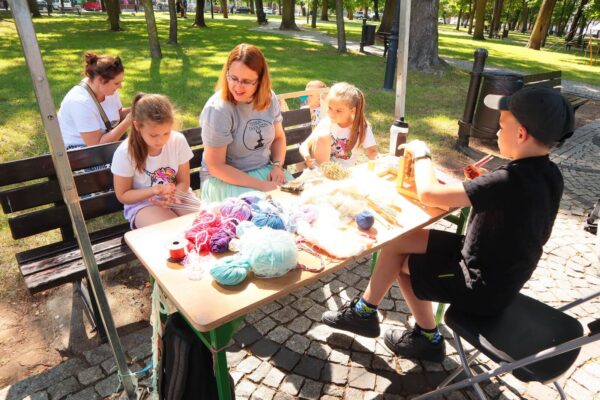 IMG_0631 warsztaty tkackie dzieci pod okiem edukatorki wyplatają bransoletki siedząc przy stole, na którym leżą materiały, chłopiec pracuje na minikrośnie tkackim