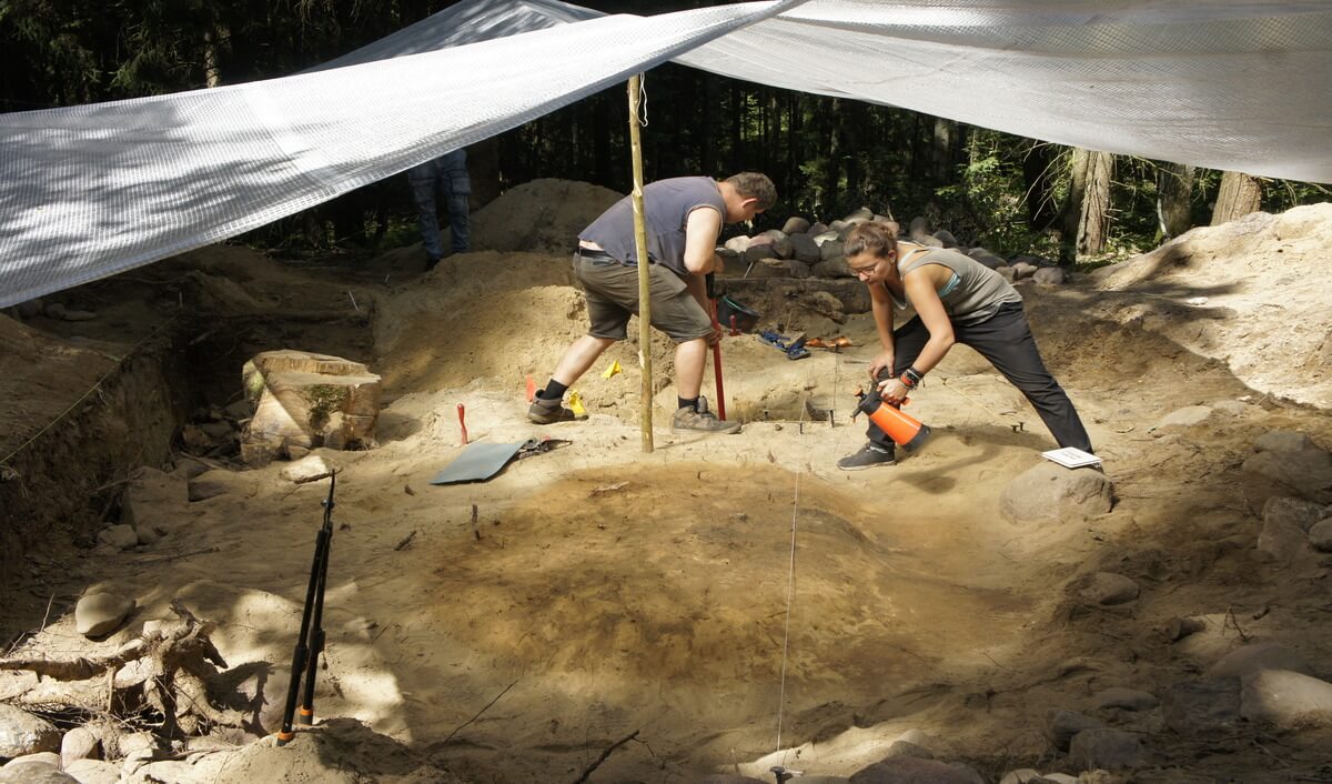 Ujęcie na wykopie, gdy Piotrek schyla się wycinając sekatorem korzeń, a Iwona schyla się kwazarując plan obiektu do zdjęcia