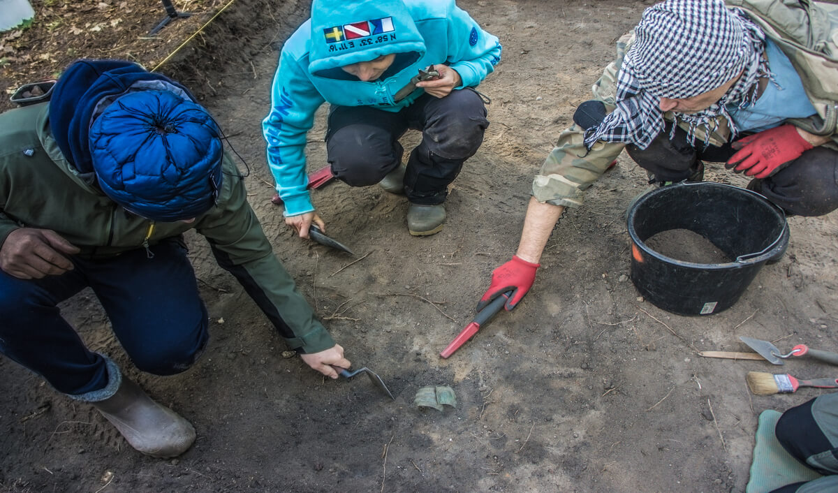 Zdjęcie bransolet in situ, gdy dwie osoby doczyszają przestrzeń wokół zabytku, a jedna sprawdza ziemię Pinpointerem