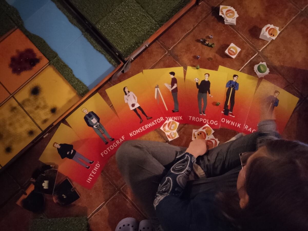 Widok od góry na siedzącą Monikę, a przed nią rozłożone są karty do gry planszowej o wykopaliskach