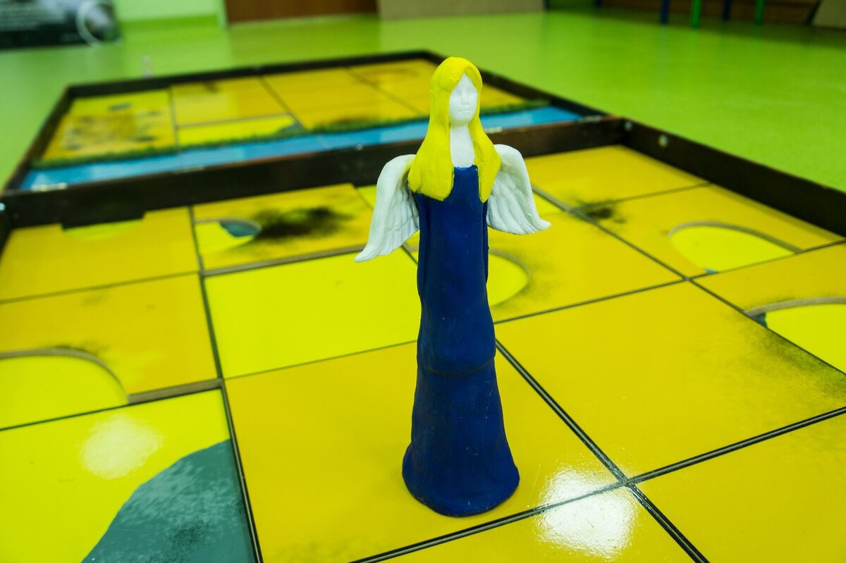 Zbliżenie na planszę, na której stoi zrobiony z masy glinianej lub solnej aniołek z małymi skrzydłami, ma żółte długie włosy i niebieska suknię