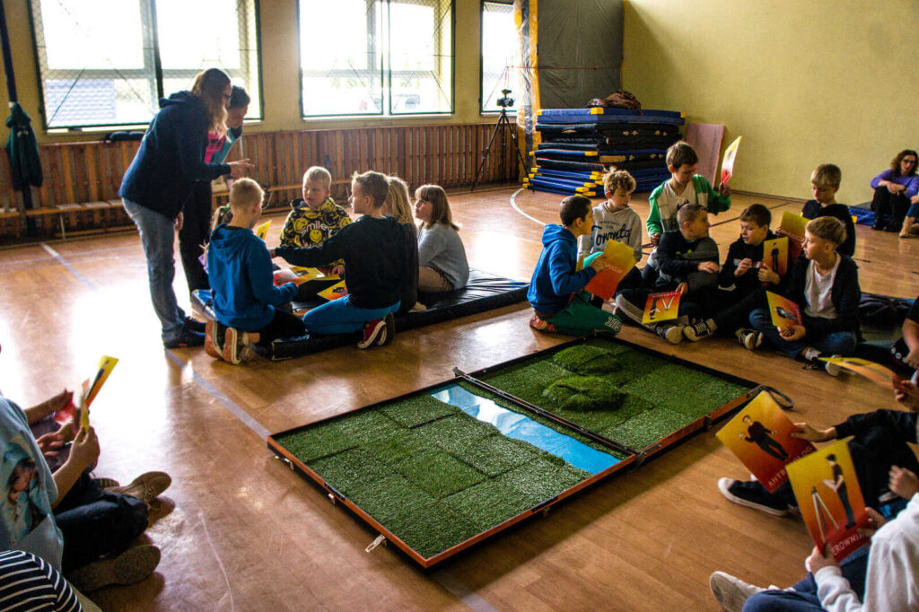 Uczniowie siedzą w grupkach na podłodze w sali sportowej i słuchają zasad gry.