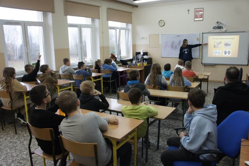 Uczniowie słuchają wykładu, a eduktorka opowiada o tym, co odkrywa archeolog.