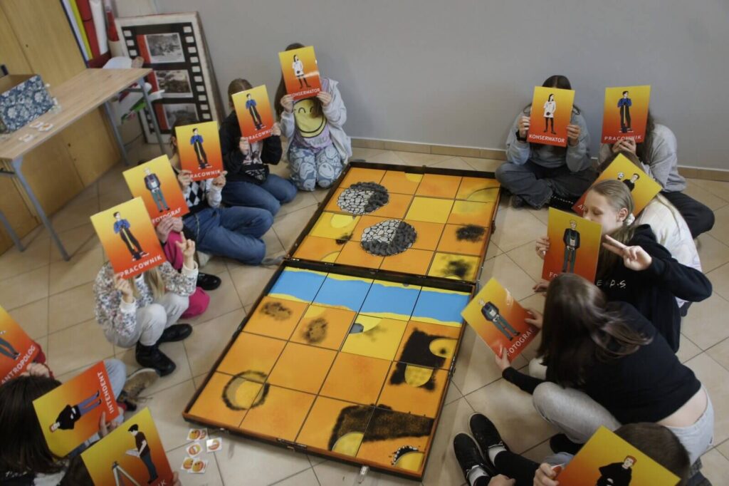 Plansza odkryta, gra skończona, gracze pozują z kartami postaci zasłaniając nimi twarze.