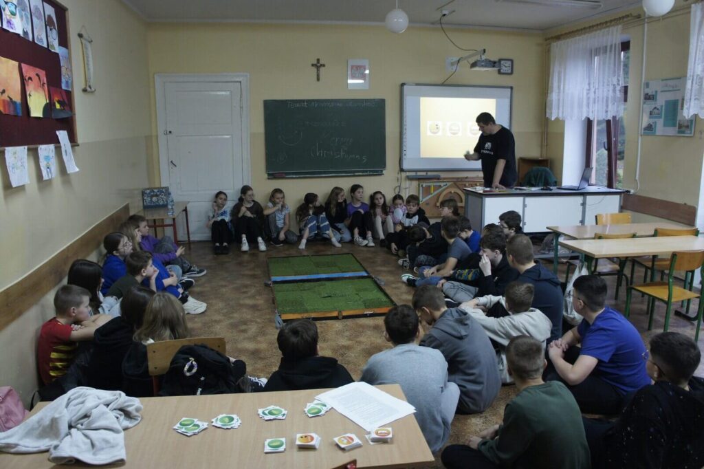 Uczniowie słuchają wprowadzenia do gry siedząc na podłodze wokół rozłożonej planszy.