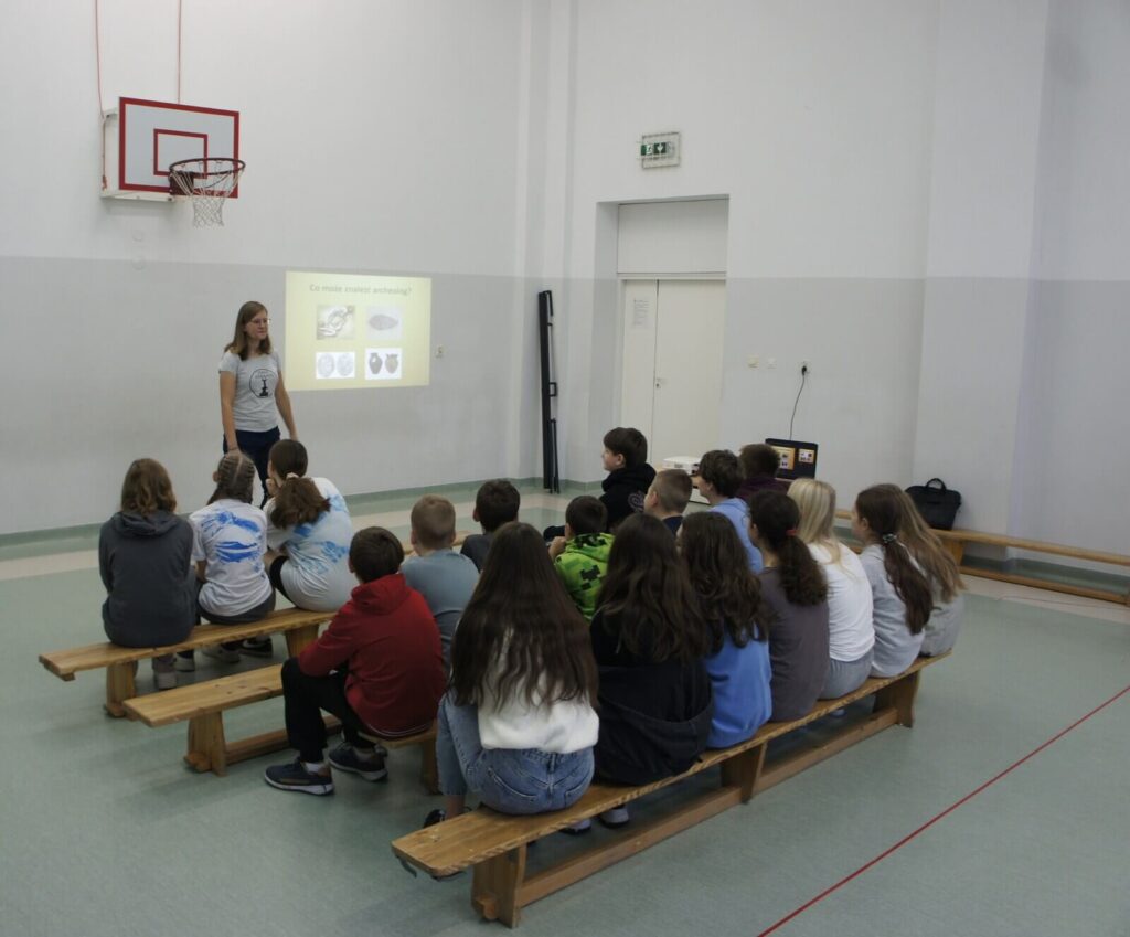 Uczniowie siedząc na ławkach w sali sportowej słuchają wykładu edukatorki.