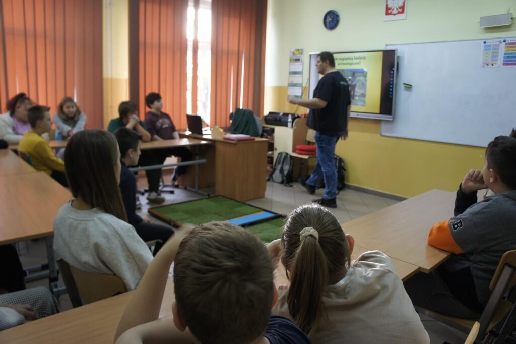 Edukator prowadzi wykład-wstęp do gry, uczniowie słuchają siedząc w ławkach.