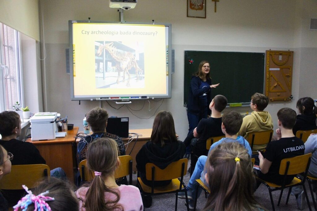 Uczniowie słuchają wykładu, a na ekranie wyświetla się pytanie" Czy archeolog bada dinozaury?