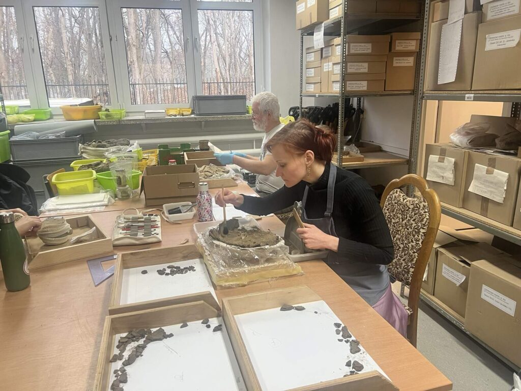 Na pierwszym planie Vini eksploruje urnę, dalej Paweł skleja fragmenty naczynia ceramicznego, na stole widać wiele sprzętów do oczyszczania i sklejania naczyń