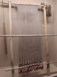 Drewniane krosno pionowe: na dwóch drążkach pionowych leżą drążki poziome, a do nich przymocowane są nici osnowy, które napinane są przez ciężarki okrągłe u dołu, z boku mamy nici wątku do tkania