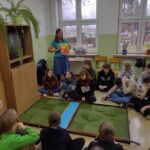 Gracze siedząc wokół planszy słuchają edukatorki.