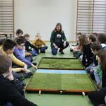 Edukatorka tłumaczy zasady gry, wszyscy siedzą wokół planszy jeszcze zakrytej.