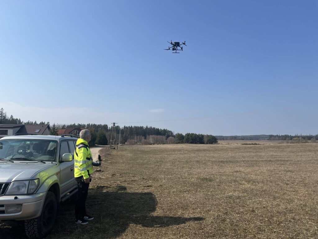 Dron unosi się w powietrzu, a z lewej stoi obok samochodu operator z pilotem w ręku.