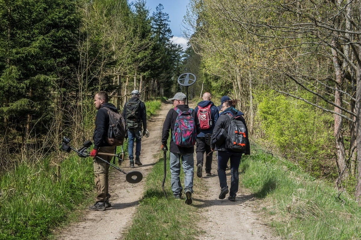 Widzimy sześć osób skierowanych do nas plecami. Idą leśną drogą trzymając wykrywacze.