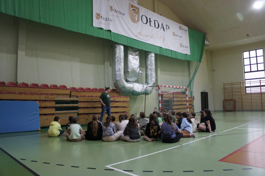 Gracze siedzą w sali gimnastycznej i słuchają edukatora