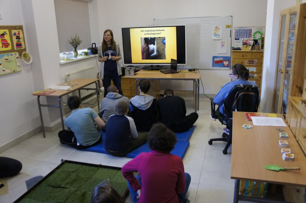 Edukatorka tłumaczy zasady uczniom siedzącym na podłodze przed ekranem prezentacji.