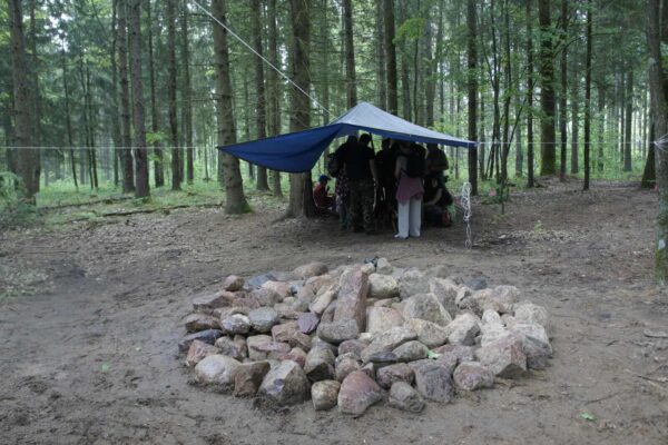 Zrekonstruowany kurhan z kamieni, w tle pod zadaszeniem z plandeki gracze i edukator.