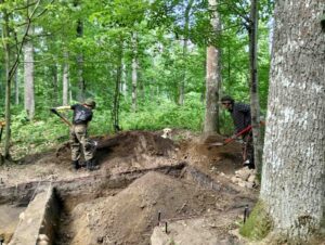 Amelia i Deborah zakopują wykop, a na hałdzie siedzi pies Mirka.