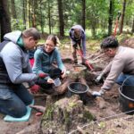 Trzy osoby kucają w wykopie pobierając próbki i eksplorując, w tle jedna pantuje wykop.
