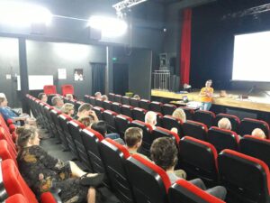 Sala kinowa, Iwona stoi pod sceną prowadząc wykład, słuchacze siedzą przed nią.