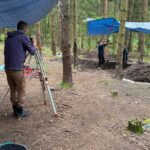 Marcin odczytuje wysokość z niwelatora, Monika trzyma łatę w miejscu pomiaru, a Natalia podnosi plandekę wiszącą nad wykopem, by ułatwić odczyt.