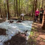 Widok na folię rozłożoną pod hałdę (ziemię z otwieranego wykopu), w tle stoi Iwona i Paweł kierujący badaniami.