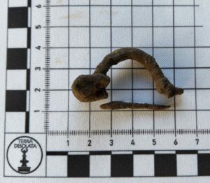 Skorodowana zapinka leży na podkładce z miarką, jest zachowana w dwóch częściach