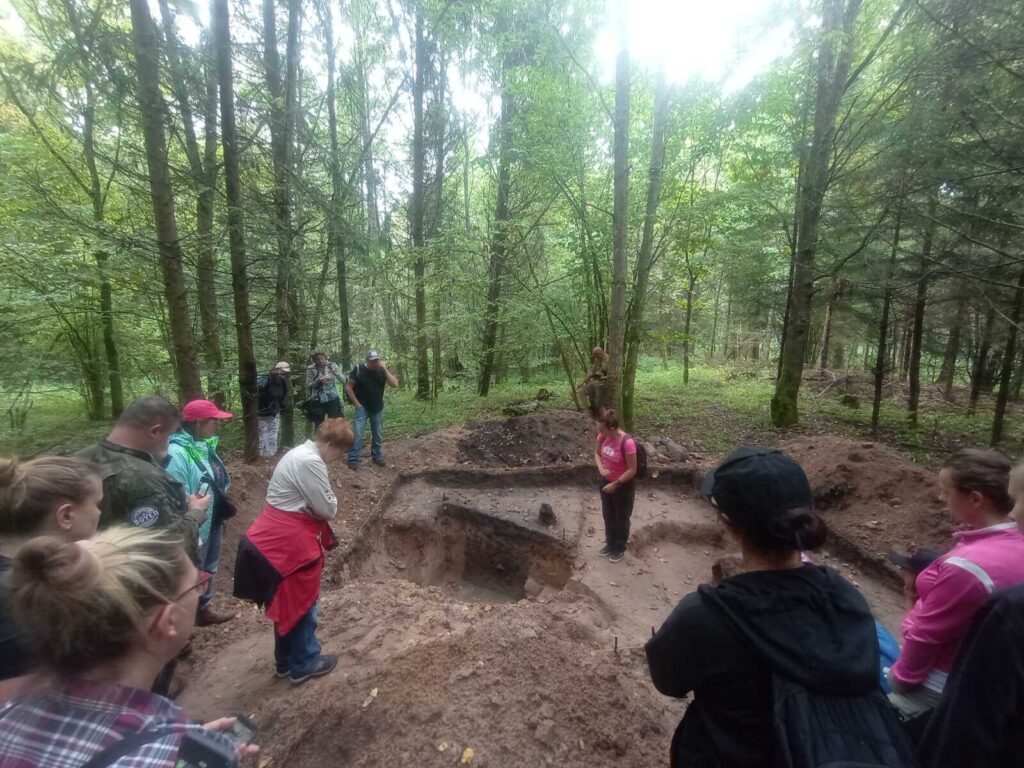 Grupa przygląda się otwartemu wykopowi archeologicznemu na osadzie bałtyjskiej. Widać profil obiektu.