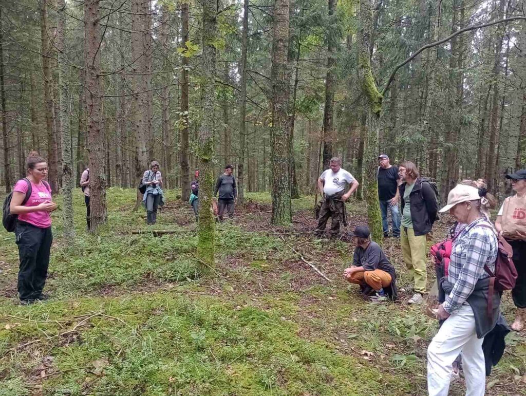 Grupa spacerowiczów stoi w lesie i słucha o cmentarzysku kurhanowym, na którym stoi...