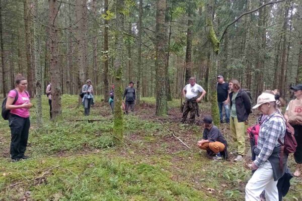 IMG_20250906_132337026_MFNR_Easy-Resize.com Grupa spacerowiczów stoi w lesie i słucha o cmentarzysku kurhanowym, na którym stoi...