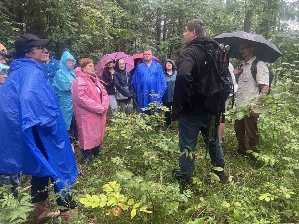 Pada deszcz. Grupa ubrana w płaszcze przeciwdeszczowe słucha edukatora opowiadającego o Jaćwingach i ich grodzisku, na którym grupa stoi.