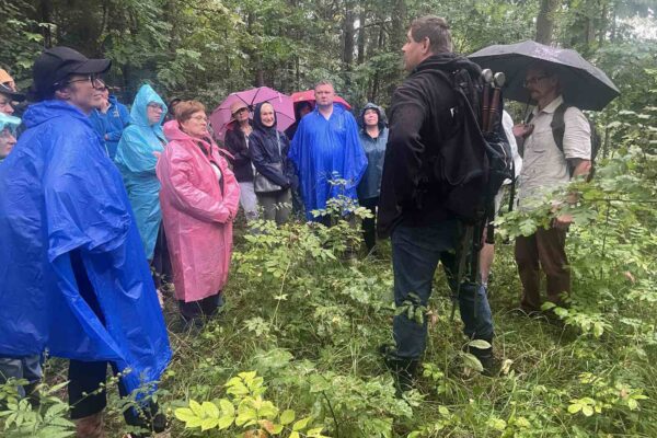 IMG_4937_Easy-Resize.com Pada deszcz. Grupa ubrana w płaszcze przeciwdeszczowe słucha edukatora opowiadającego o Jaćwingach i ich grodzisku, na którym grupa stoi.