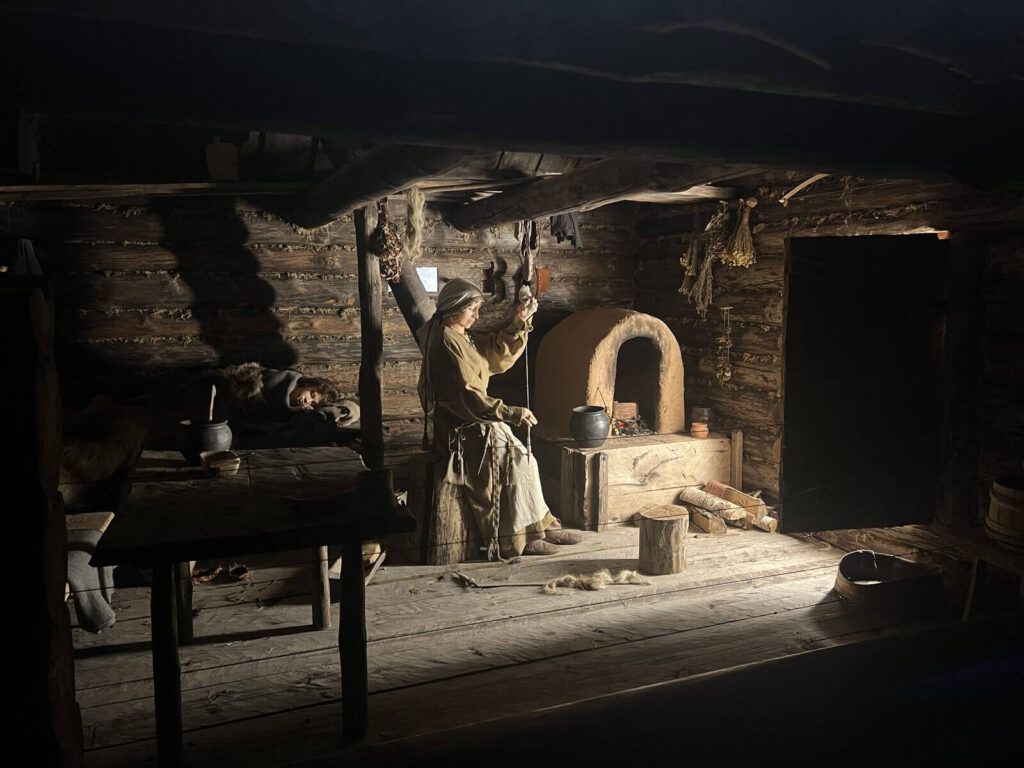 Widok na wnętrze rekonstrukcji chaty bałtyjskiej, kobieta siedzi przy glinianym piecu