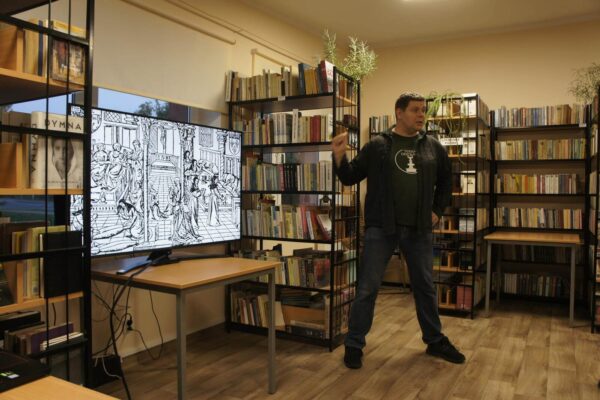 Piotr stoi na tle bibliotecznych regałów i obok ekranu z prezentacją