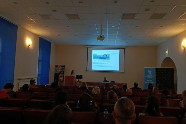 Kolejny widok z końca sali na ekran z prezentacją i z lewej przemawia Iwona