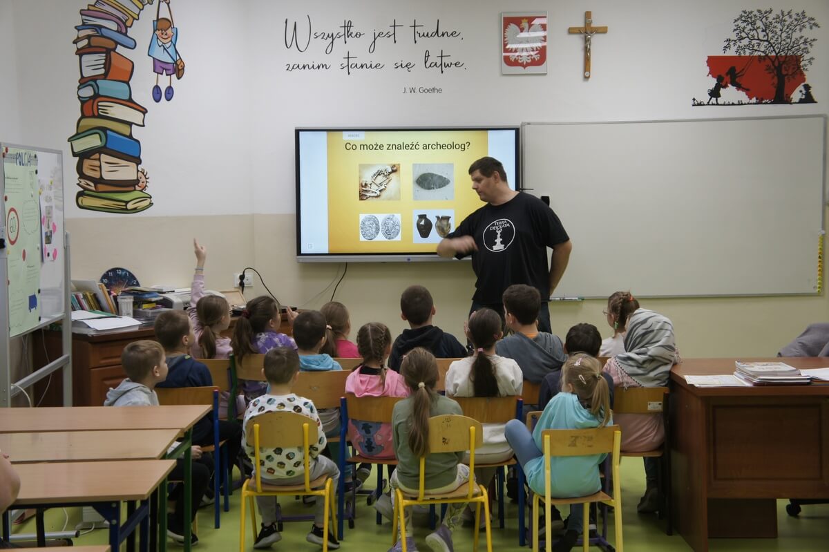 Piotrek opowiada o pracy archeologa, w tle prezentacja wyświetlana na telebimie. Przed nim siedzi grupa uczniów.