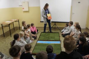 Monika stojąc nad planszą i siedzącymi wokół uczniami omawia zasady gry.