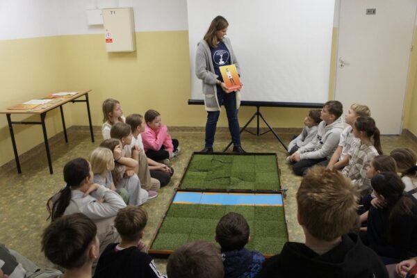 Monika stojąc nad planszą i siedzącymi wokół uczniami omawia zasady gry.