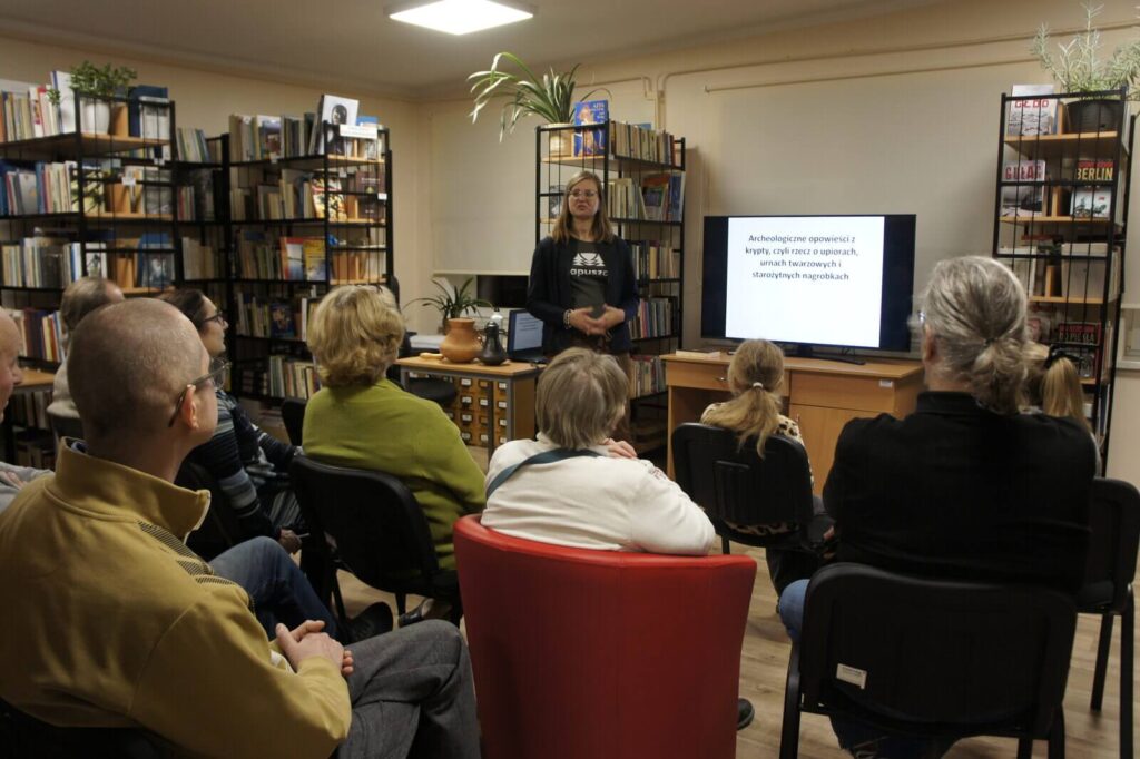 W głębi Monika stojąc prowadzi wykład. Obok niej wyświetlana jest na monitorze prezentacja. Na pierwszym planie widoczni tyłem siedzący słuchacze.
