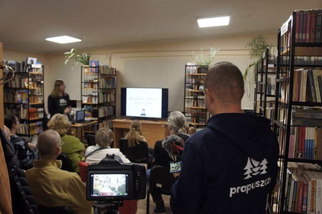 Na pierwszym planie kamera i stojący obok Bartek nagrywający wykład. W tle słuchacze i Monika prowadząca wykład.