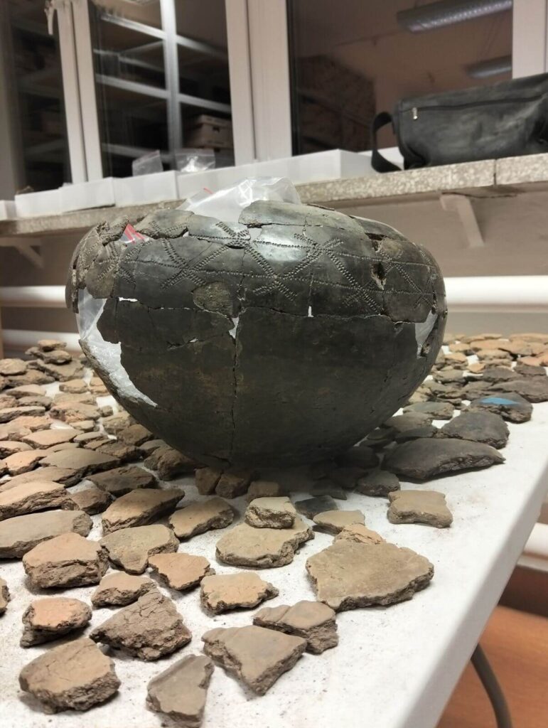 Sklejona urna stoi na fragmentach ceramiki leżących na stole.