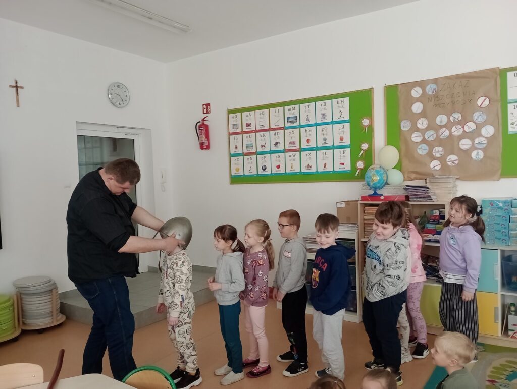 Dzieci stoją w kolejce, a Piotrek zakłada każdemu po kolei rekonstrukcję hełmu średniowiecznego.