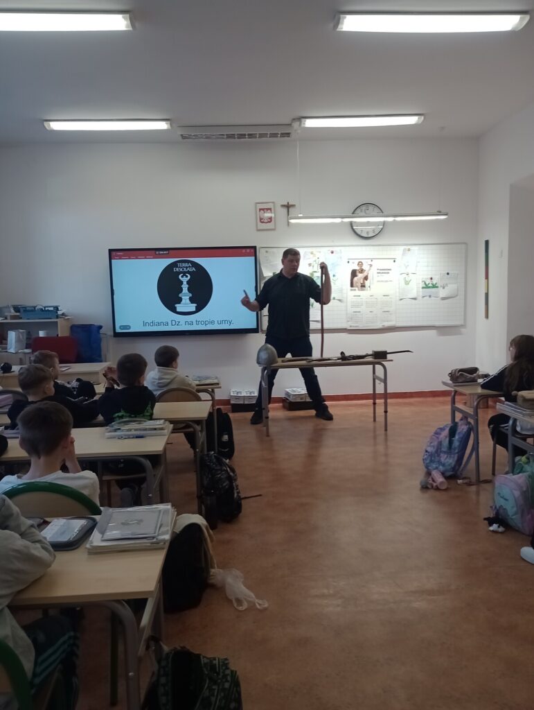Piotrek omawia temat zajęć stojąc przy prezentacji, dzieci siedzą w ławkach.