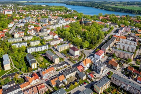 Widok z drona na zadrzewiony rynek i ulice i budynki wokół, w tle Jezioro Oleckie Wielkie