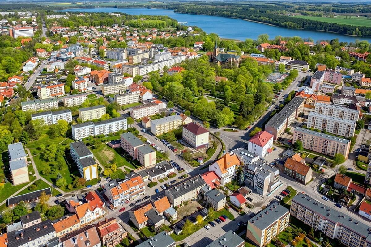 Widok z drona na zadrzewiony rynek i ulice i budynki wokół, w tle Jezioro Oleckie Wielkie