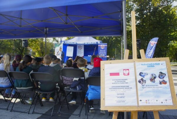 Na pierwszym tle plakaty wydarzenia i dotyczący dofinansowania, w tle publiczność słuchająca wykładu.