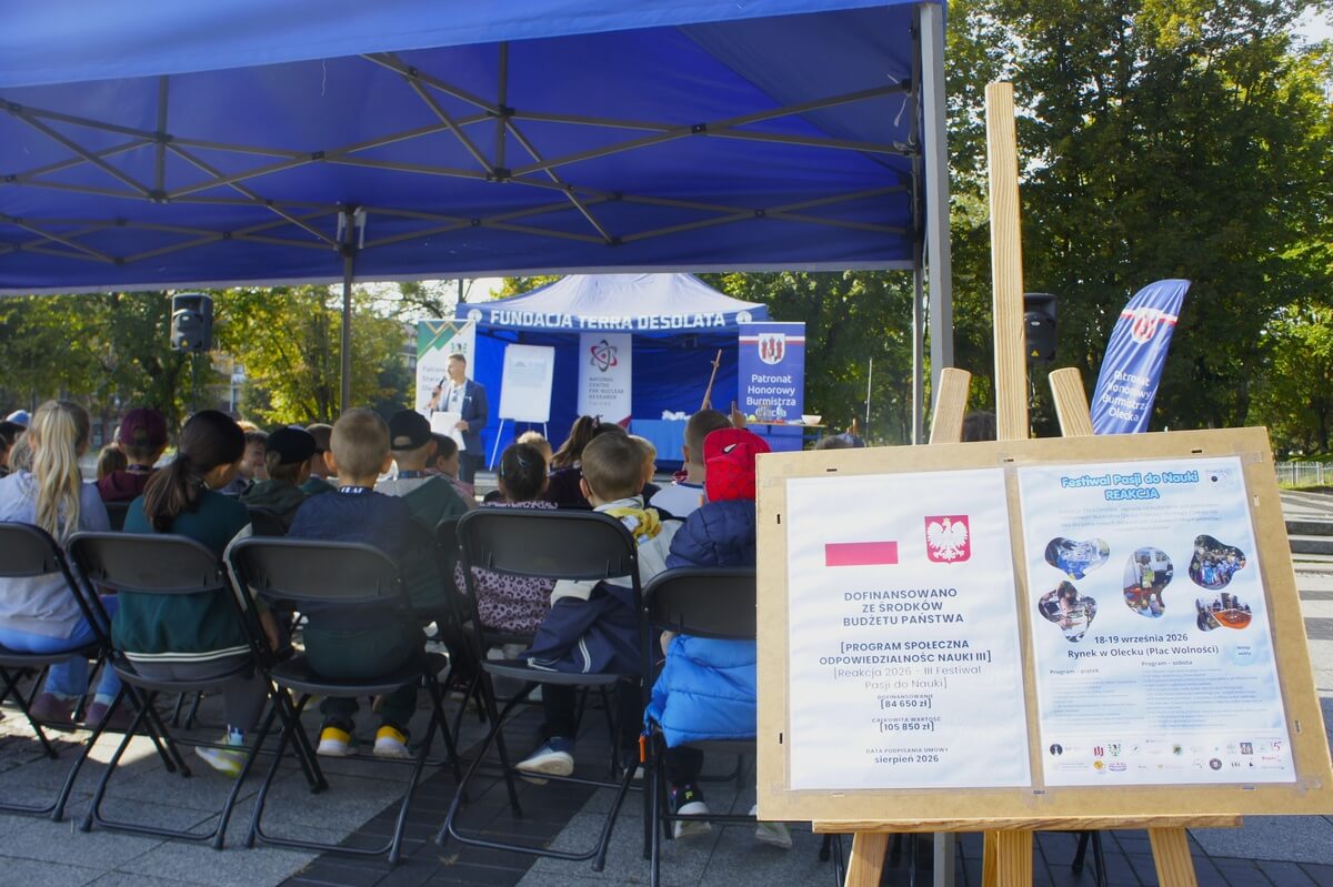 Na pierwszym tle plakaty wydarzenia i dotyczący dofinansowania, w tle publiczność słuchająca wykładu.